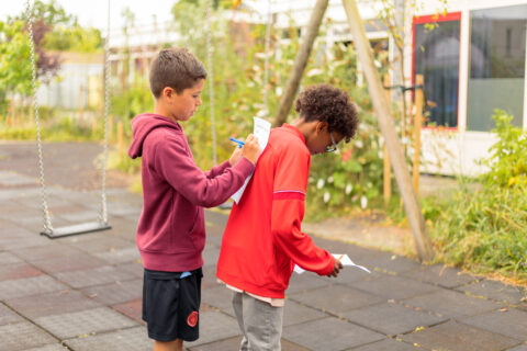 Twee jongens buiten, spelend met pen en papier. Eén schrijft op de rug van de ander.