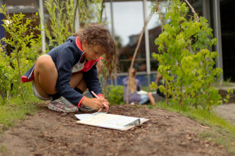 Meisje schrijft buiten op een klembord, omgeven door planten, met kinderen op de achtergrond.