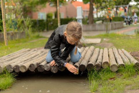 Meisje in leren jas schept water onder houten brug op een grasveld in een park.