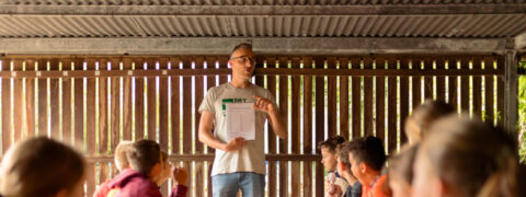 Een man geeft uitleg aan kinderen aan een lange tafel in een houten ruimte.