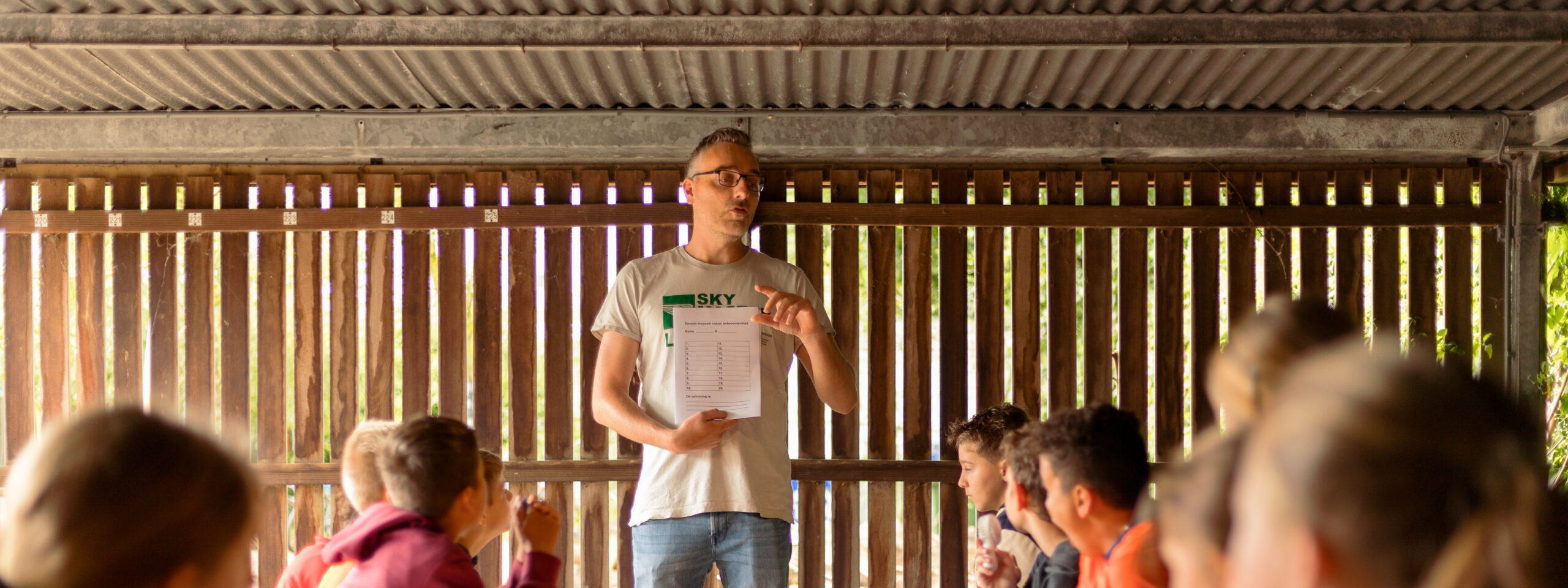 Een man geeft uitleg aan kinderen aan een lange tafel in een houten ruimte.