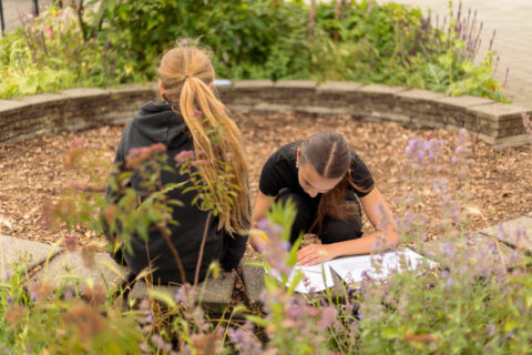 Twee kinderen buiten, zittend in een cirkel, bezig met papierwerk, omringd door planten.