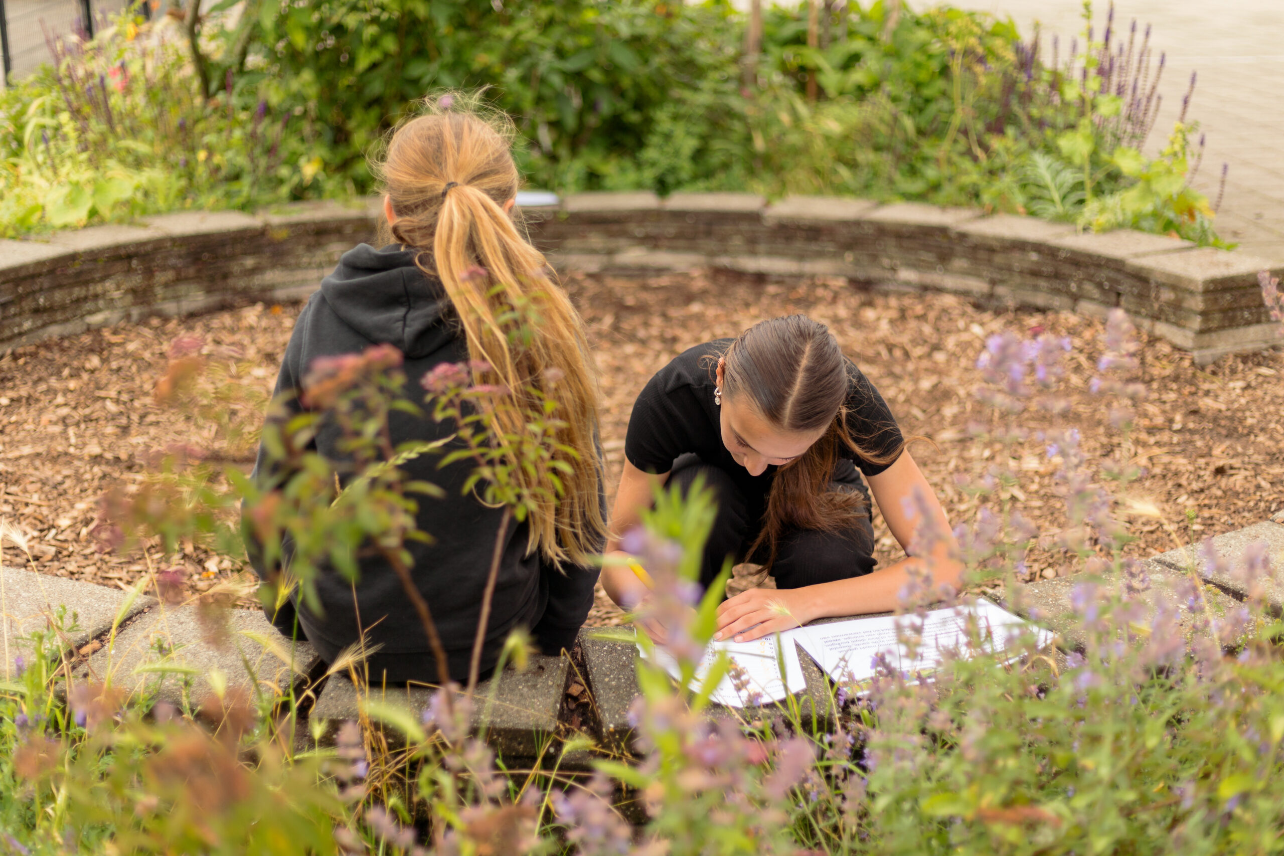 Twee kinderen buiten, zittend in een cirkel, bezig met papierwerk, omringd door planten.