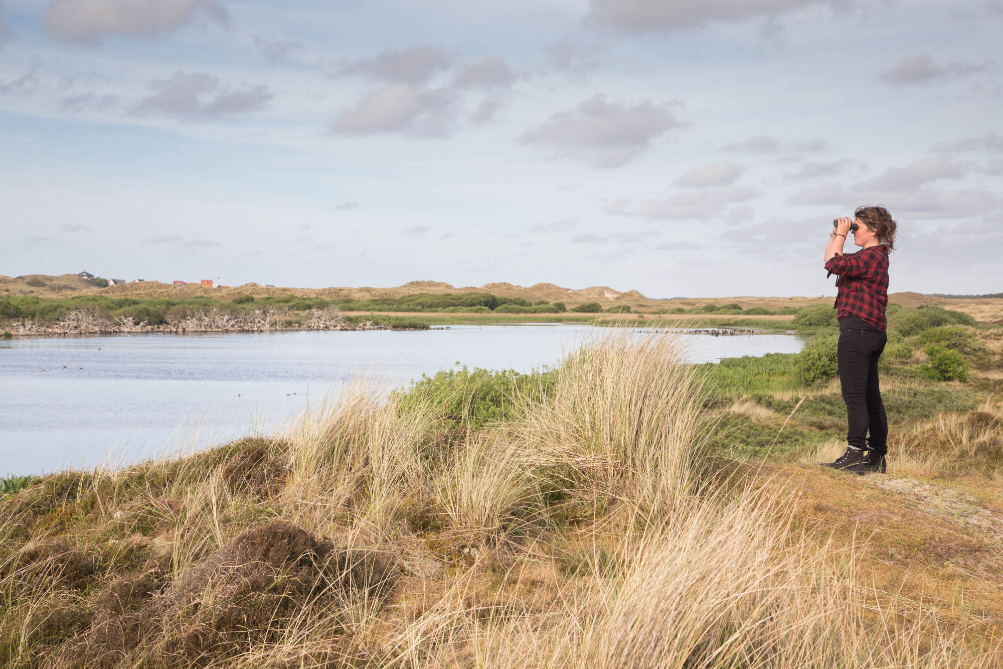 Persoon kijkt met verrekijker naar meer in duinlandschap onder bewolkte lucht.