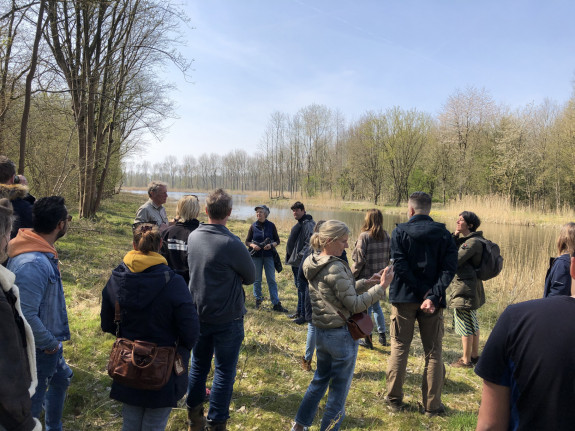 Groep mensen bij een bosrand luistert naar een spreker, met een meer en bomen op de achtergrond.