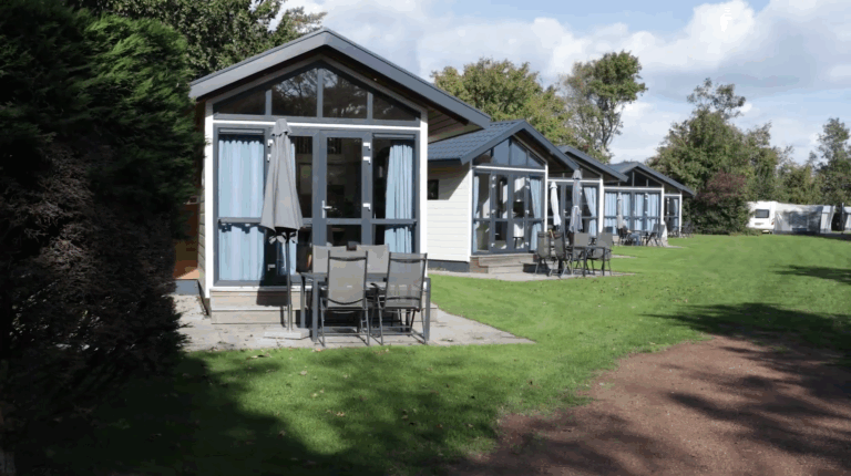 Rij vakantiehuisjes met tuinmeubels en parasols op grasveld in zonnige omgeving.