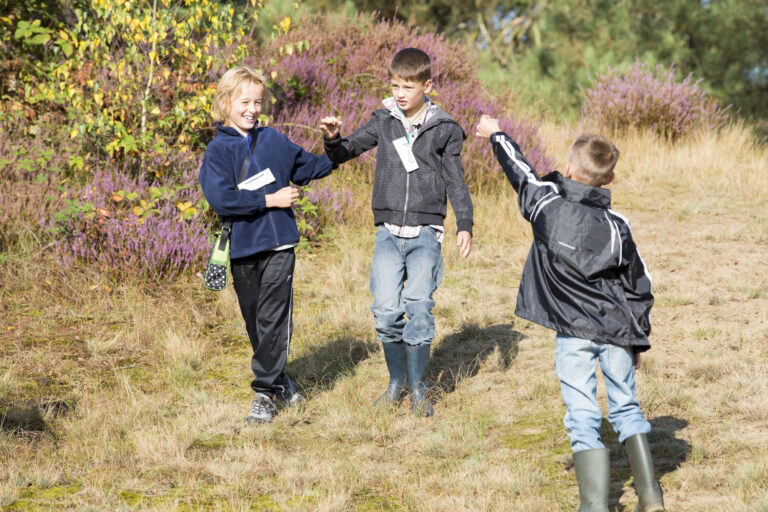 Drie kinderen lopen en spelen buiten in een grasrijke omgeving met kleurrijke struiken.