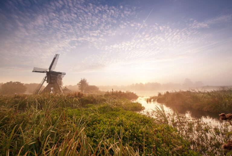Ochtendzicht op een oude windmolen bij een nevelige rivier, omgeven door graslanden en blauwe lucht.