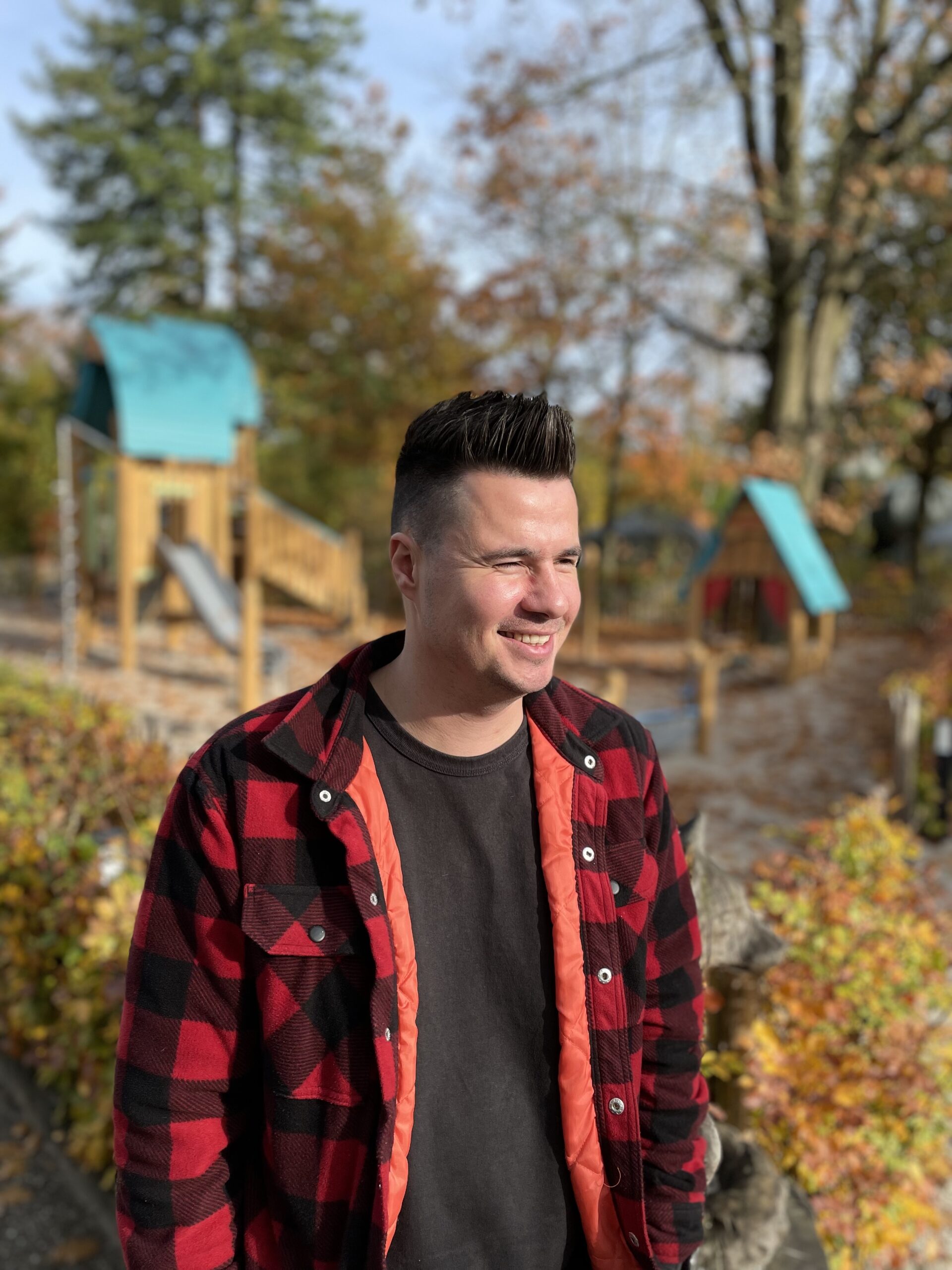 Man in rood-zwart geruite jas lacht in park met herfstbladeren en speeltoestellen.