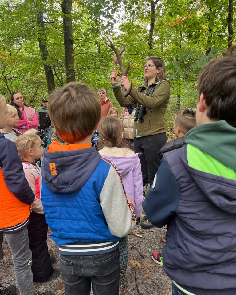 Een vrouw toont een gewei aan kinderen in een bos tijdens een outdoor educatieve activiteit.