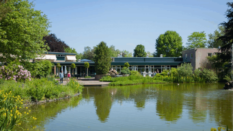 Groot gebouw met terras aan vijver, omgeven door groene planten en bomen op een zonnige dag.