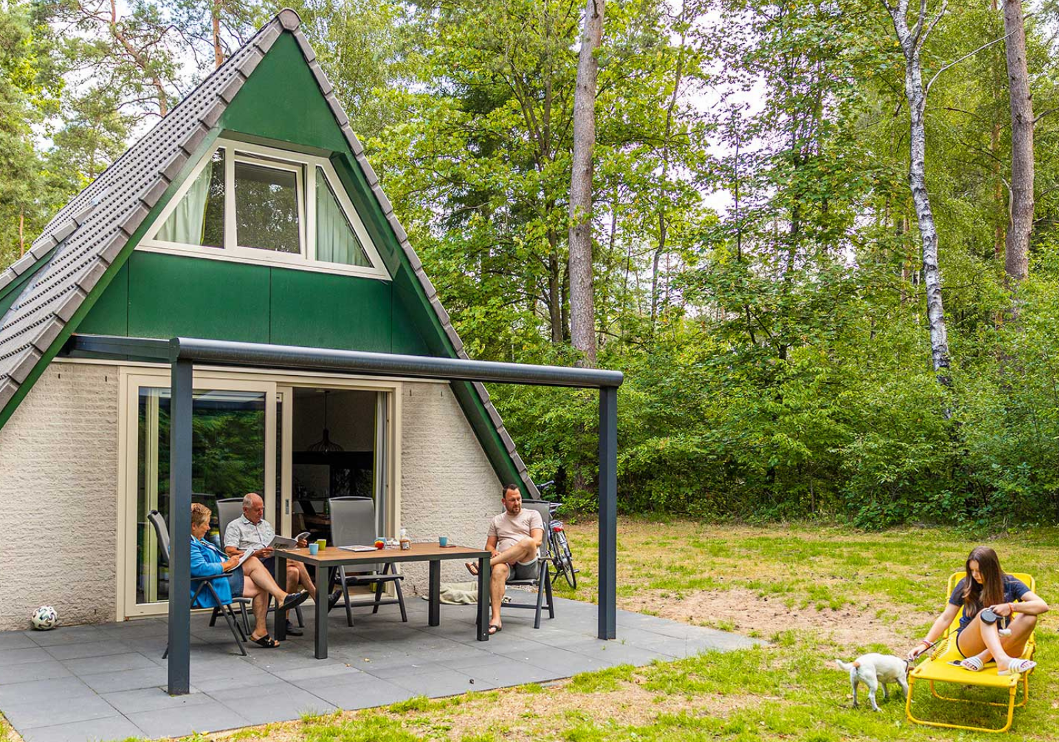 Groen huis met terras in bos; mensen zitten buiten, vrouw speelt met hond.