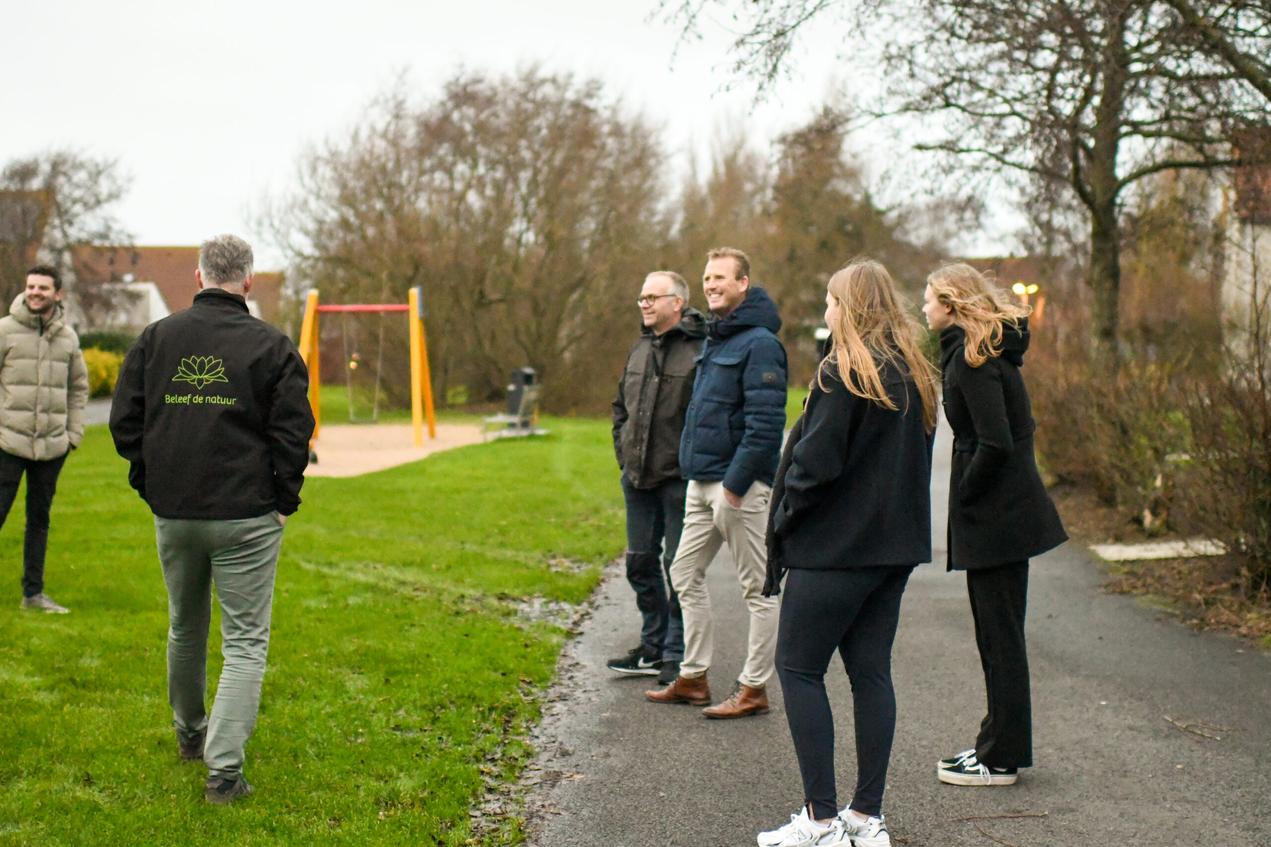 Groep mensen wandelt in park, één draagt jas met tekst "Beleef de natuur". Speeltoestel op achtergrond.