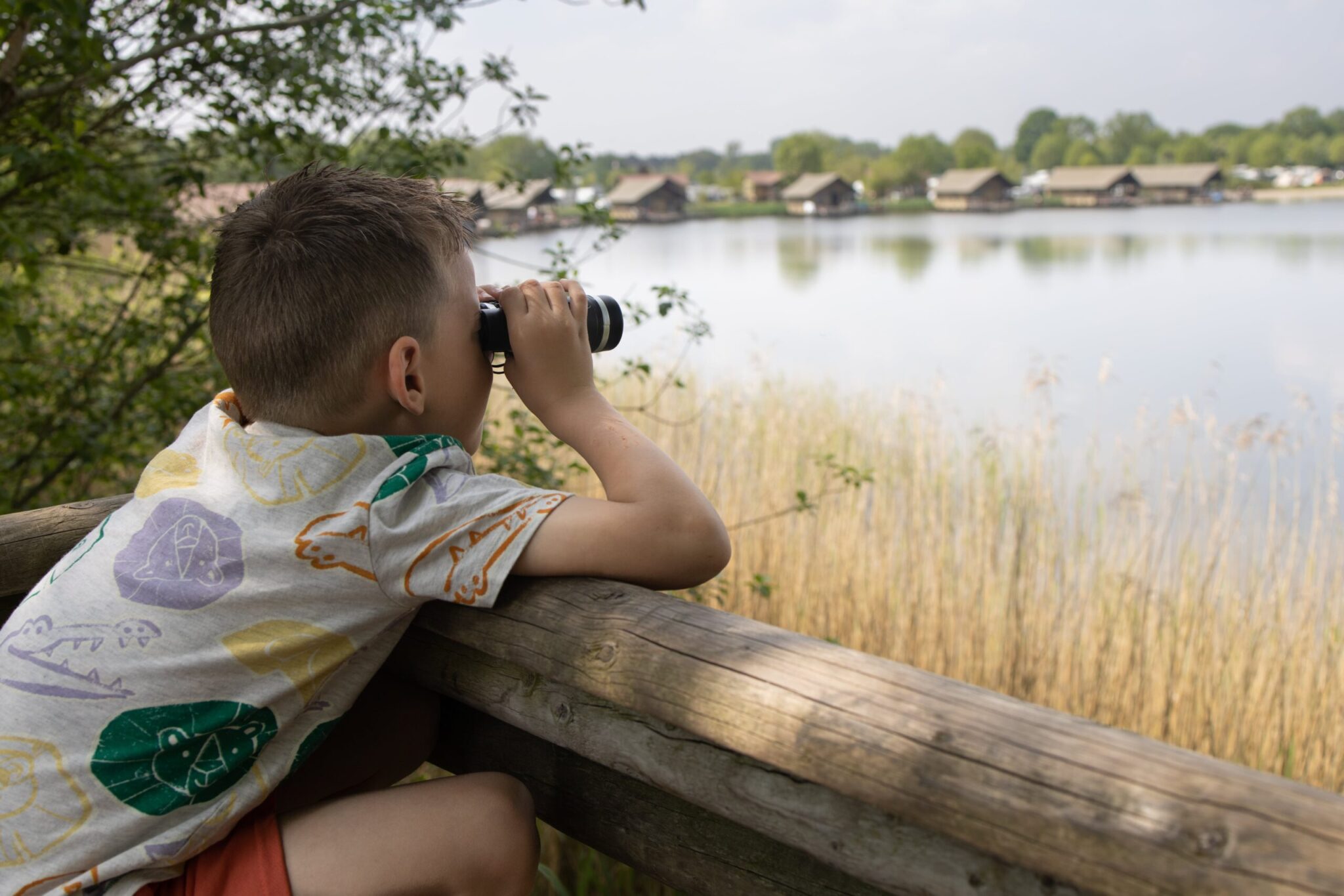 Kind kijkt met verrekijker over meer vanaf houten hek. Hutten zichtbaar aan andere kant van het water.