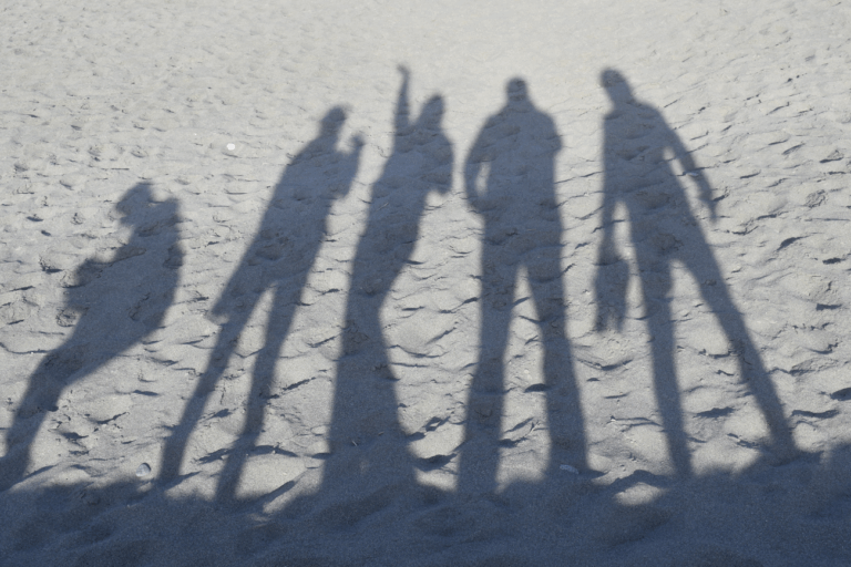 Schaduwen van vijf mensen op een zandstrand, met een persoon die gebaart.