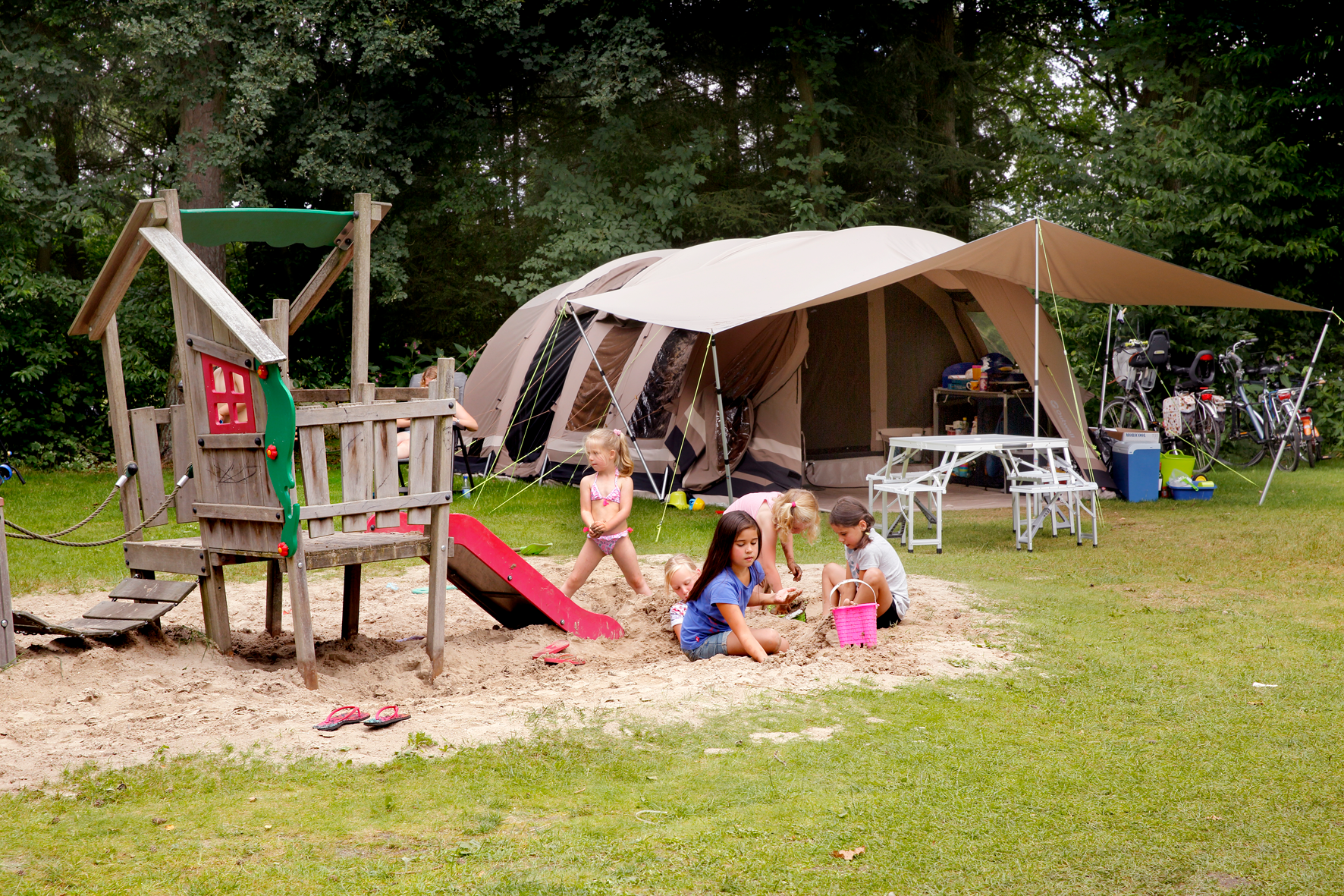 Kinderen spelen in een zandbak bij een grote tent op een camping, omringd door bos.