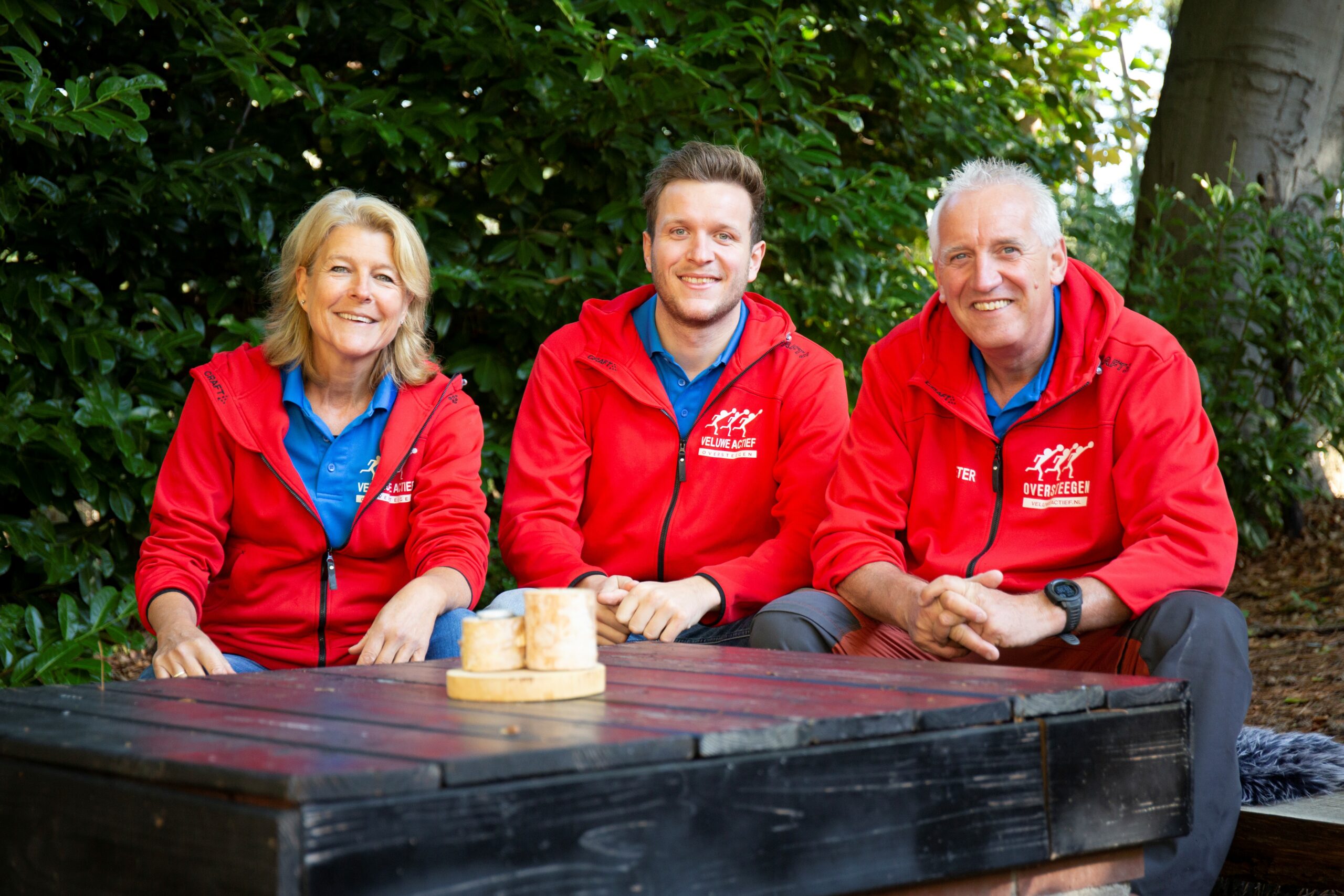 Drie mensen in rode jassen zitten buiten aan een houten tafel, met groen gebladerte op de achtergrond.