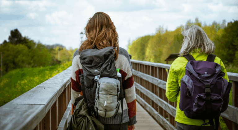 Twee personen met rugzakken wandelen over een houten brug in een bosrijke omgeving.