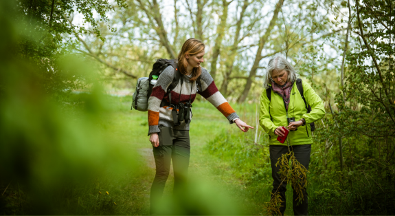 Twee vrouwen wandelen in een groen bos, al pratend en onderzoekend naar planten.