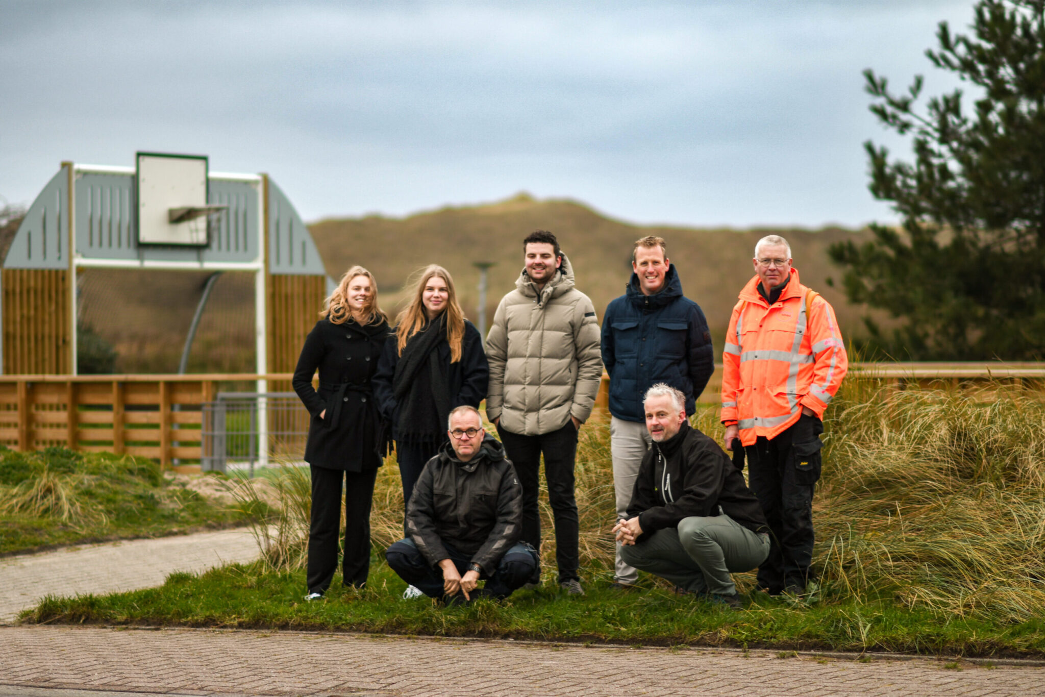 Groep van zeven mensen staat en hurkt op een pad bij een basketbalveld met gras en duinen op de achtergrond.