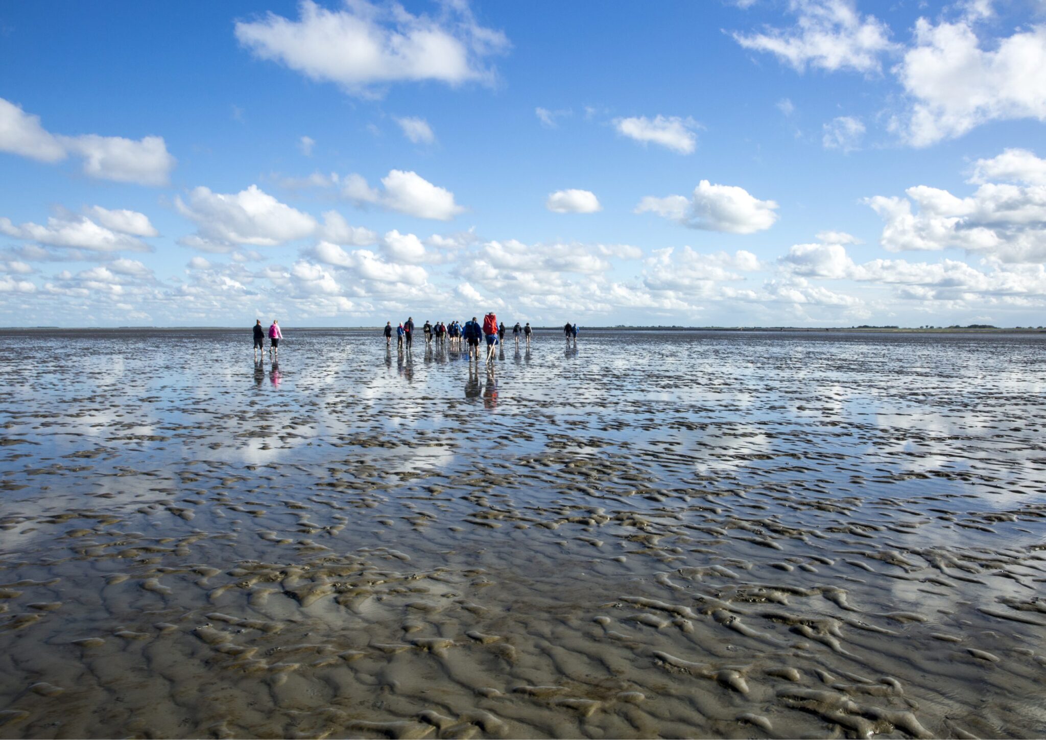 Mensen wandelen op een uitgestrekte, natte zandvlakte onder een blauwe lucht met witte wolken.