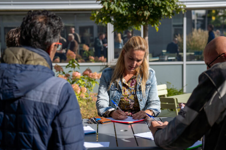 Een vrouw schrijft aan een tafel buiten, omringd door andere mensen en groene planten.