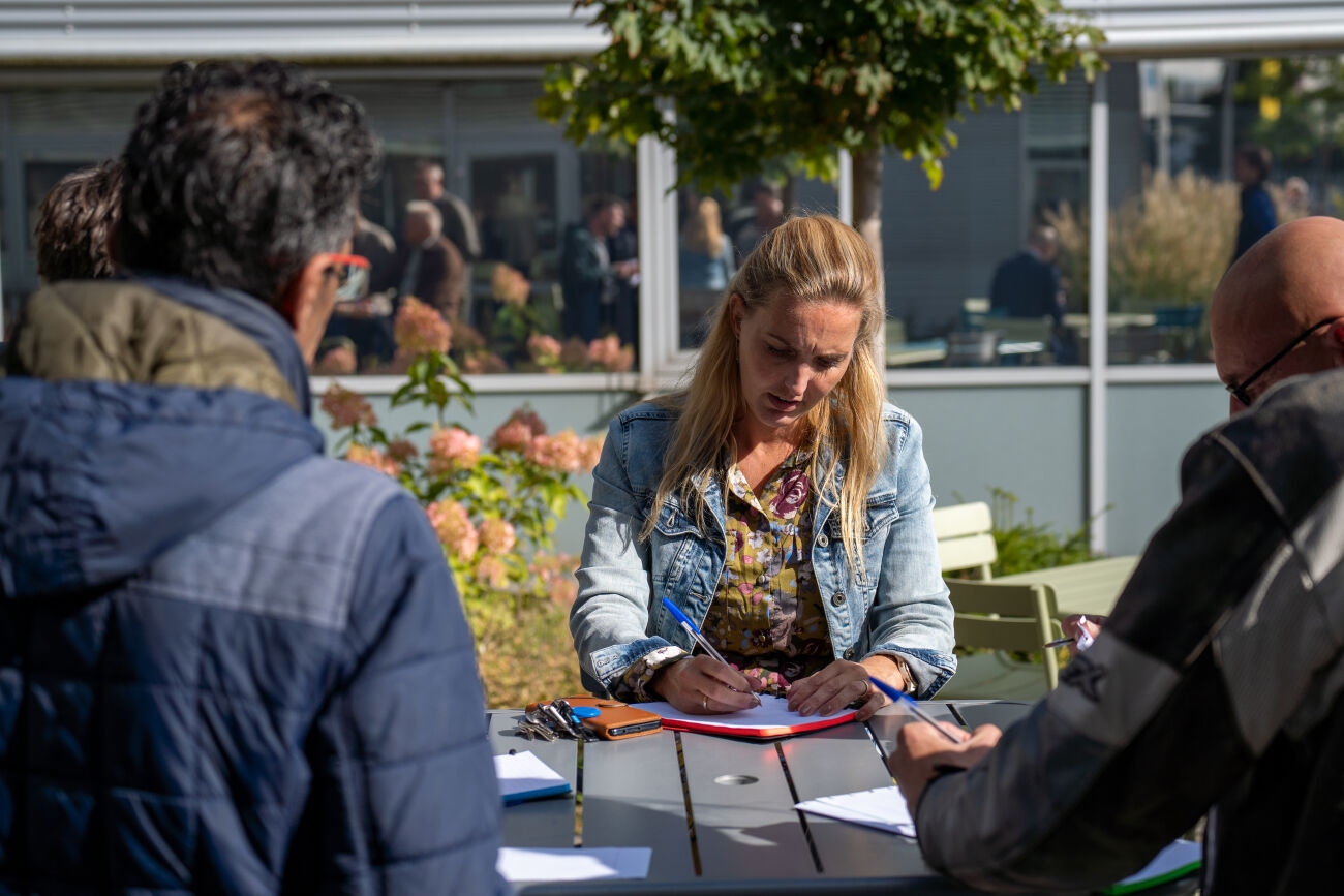Een vrouw schrijft aan een tafel buiten, omringd door andere mensen en groene planten.