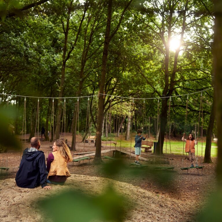 Twee mensen zitten op een heuvel in een bos, terwijl kinderen nabij op speeltoestellen spelen.