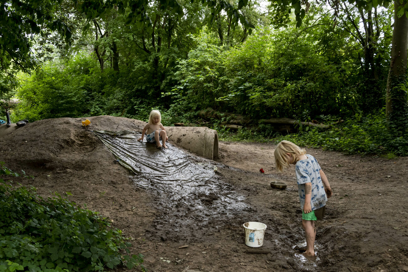 Twee kinderen spelen in de modder met een geïmproviseerde glijbaan in een bosomgeving.