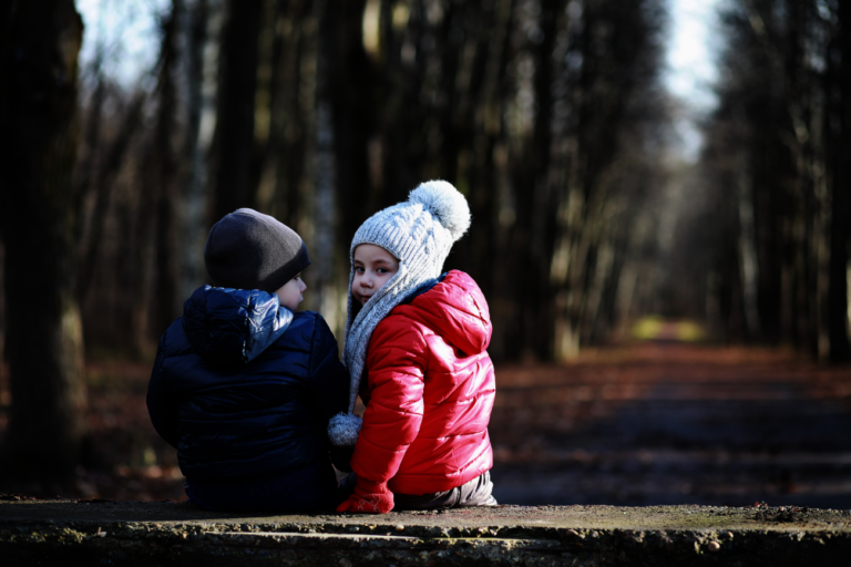 Twee kinderen in winterjassen en mutsen zitten op een bankje in een bos.
