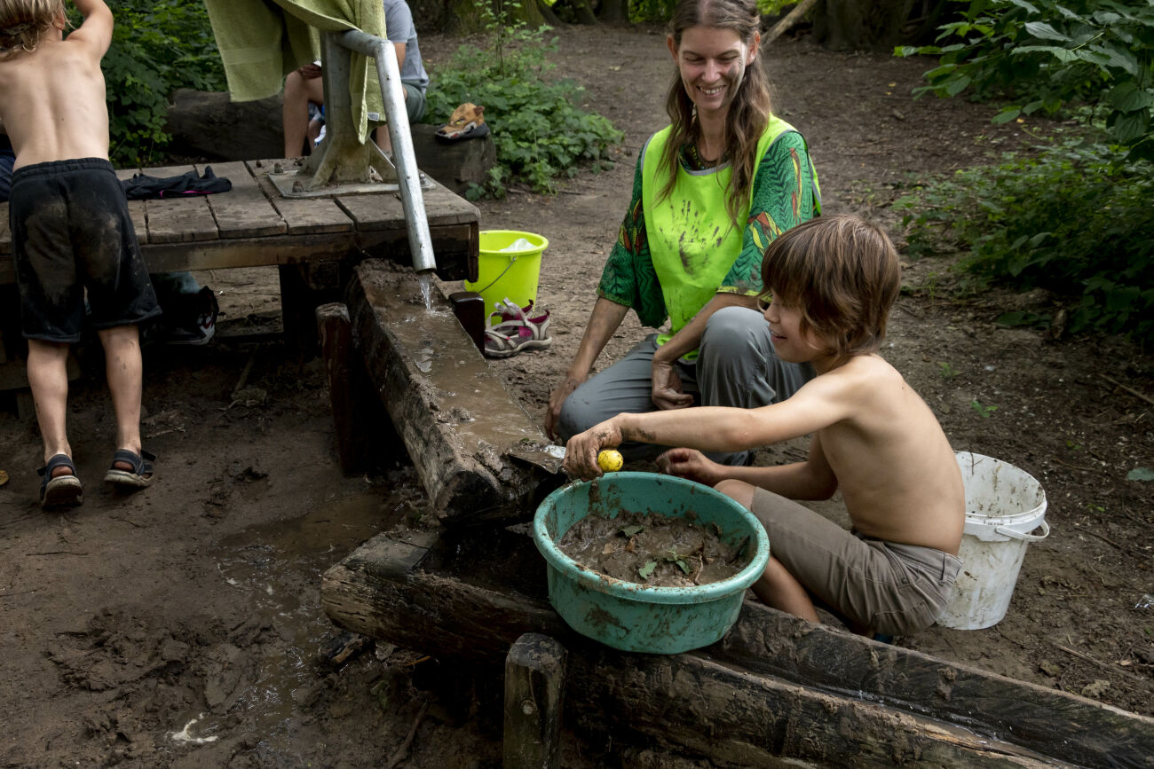 Kinderen spelen met modder en water in een bosachtige omgeving, begeleid door een lachende volwassene.