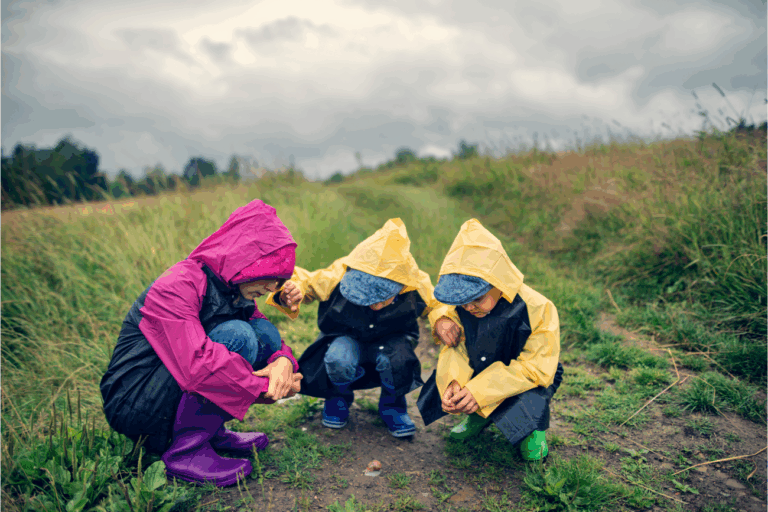 Drie kinderen in regenjassen kijken naar de grond op een pad tussen grasvelden.