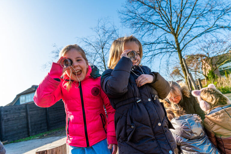 Twee meisjes buiten in jassen kijken door kartonnen kijkers, lachend in de zon.