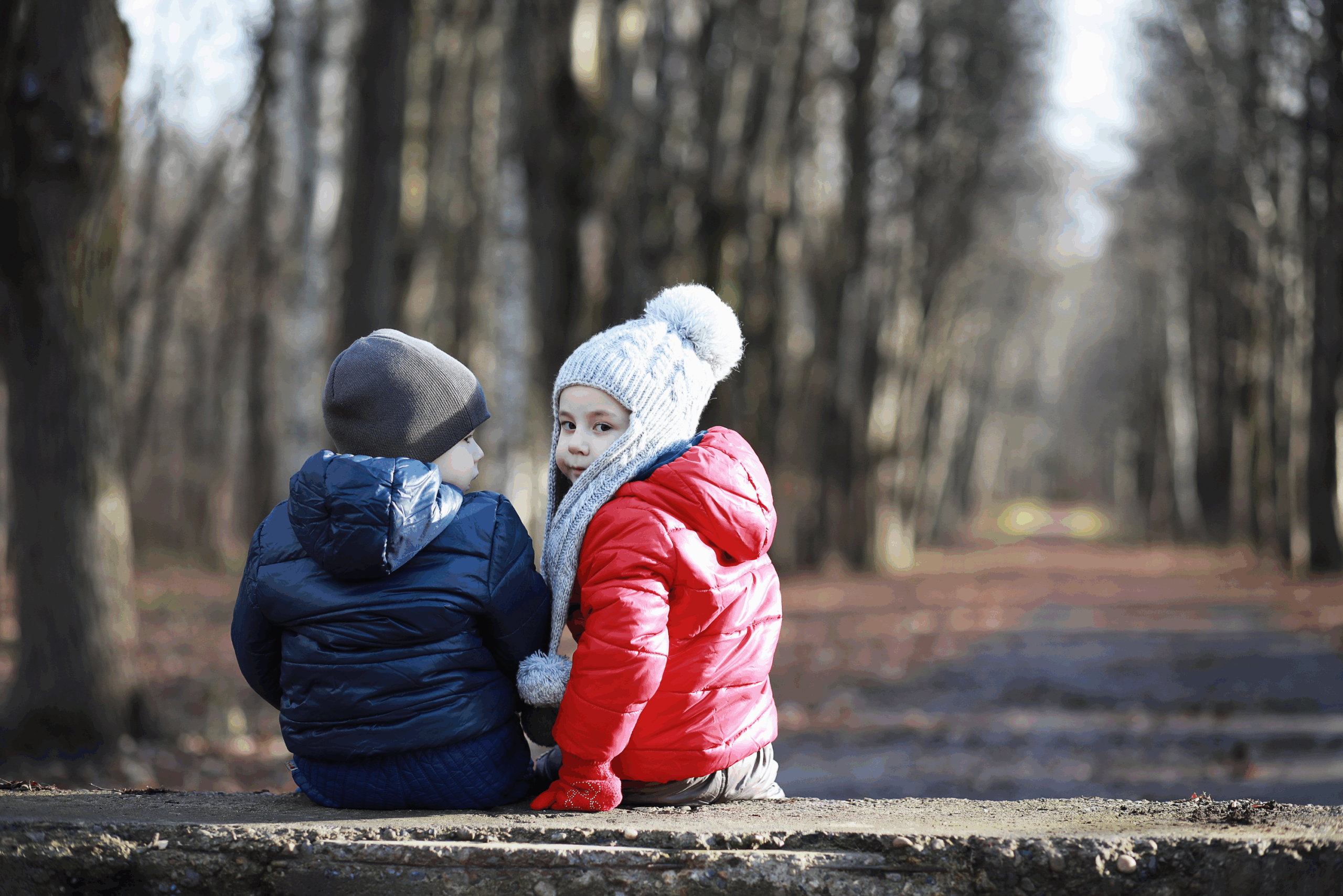 Twee kinderen in winterjassen zitten op een bank in een bosrijke omgeving.