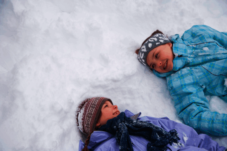 Twee kinderen spelen liggend en lachend in de sneeuw, gekleed in winterkleding.
