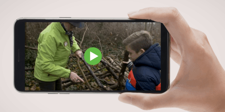 Een hand houdt een telefoon vast met een video van een kind en volwassene die takken onderzoeken.