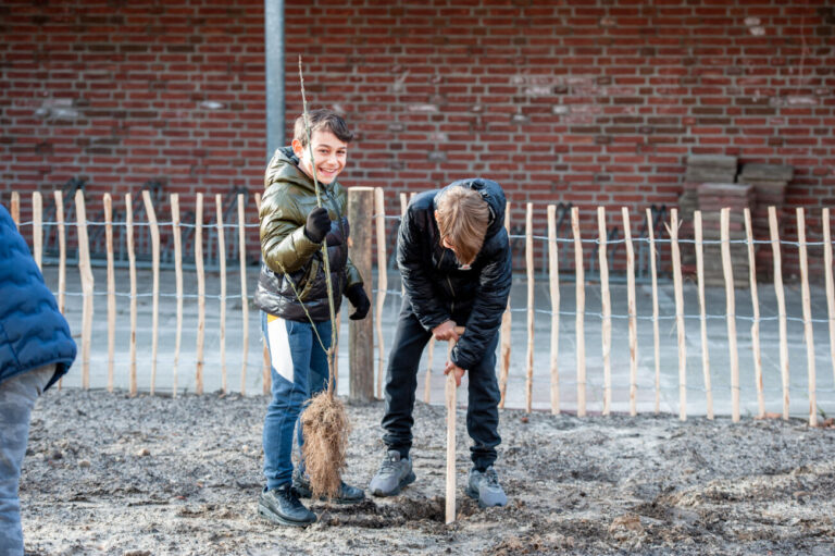 Kinderen planten een boom in een modderige tuin met een houten hek en bakstenen muur op de achtergrond.
