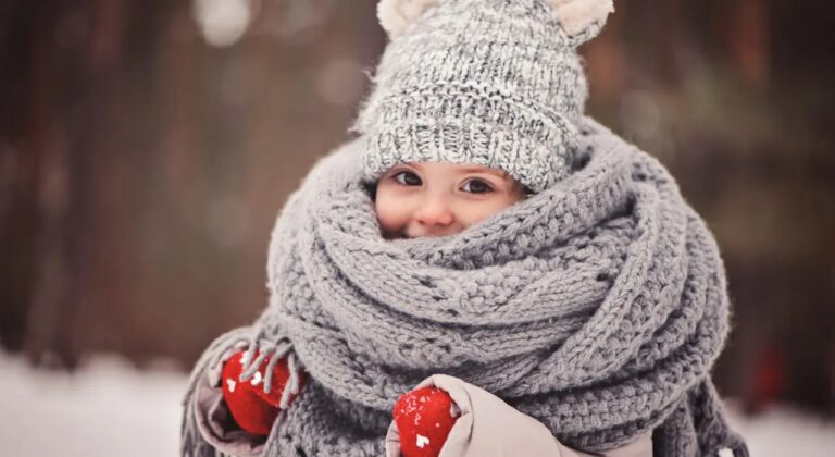 Kind ingepakt in grijze sjaal en muts met rode wanten, in een winterse omgeving.
