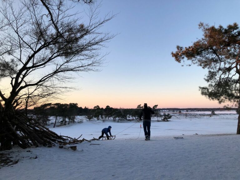 Twee mensen trekken een slee door een besneeuwd landschap bij zonsondergang.