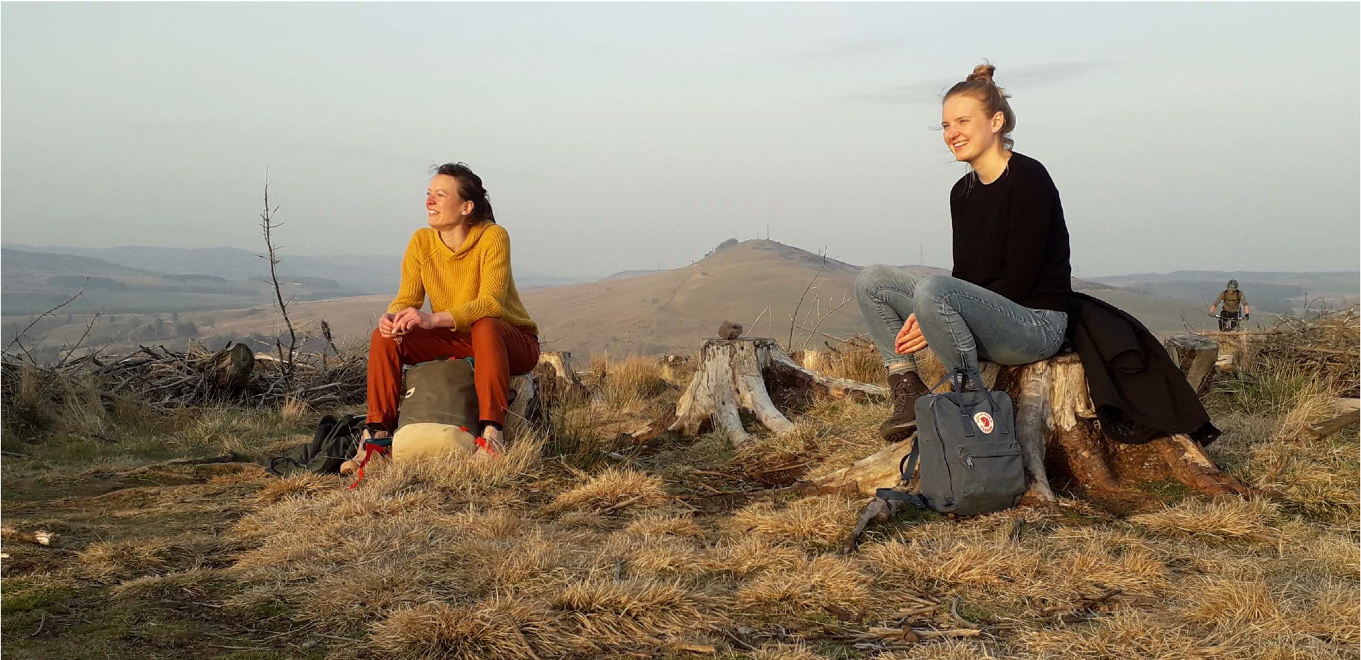 Twee vrouwen zitten op boomstronken met rugzakken in een heuvelachtig landschap bij zonsondergang.