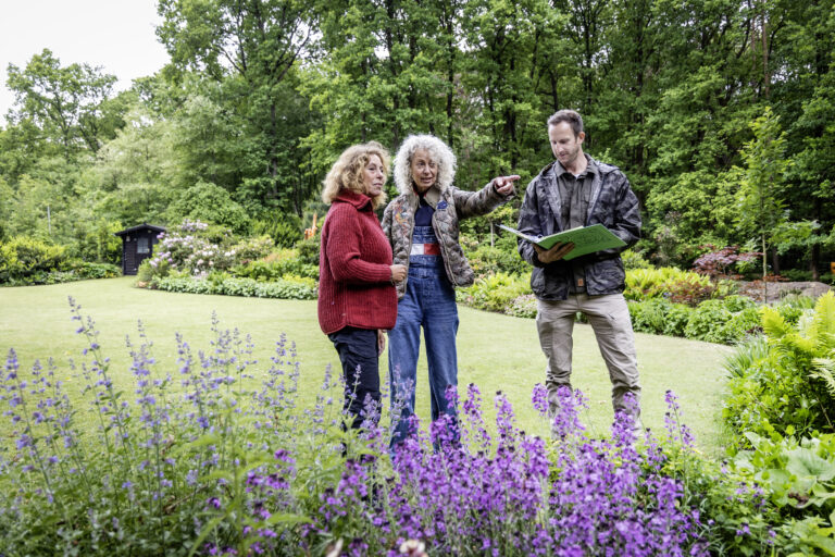 Drie mensen in een tuin, pratend en wijzend, met een boek in de hand, omringd door bloemen en bomen.