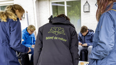 Groep mensen in blauwe jassen, één met "Beleef de natuur" logo, bezig met een activiteit buiten.