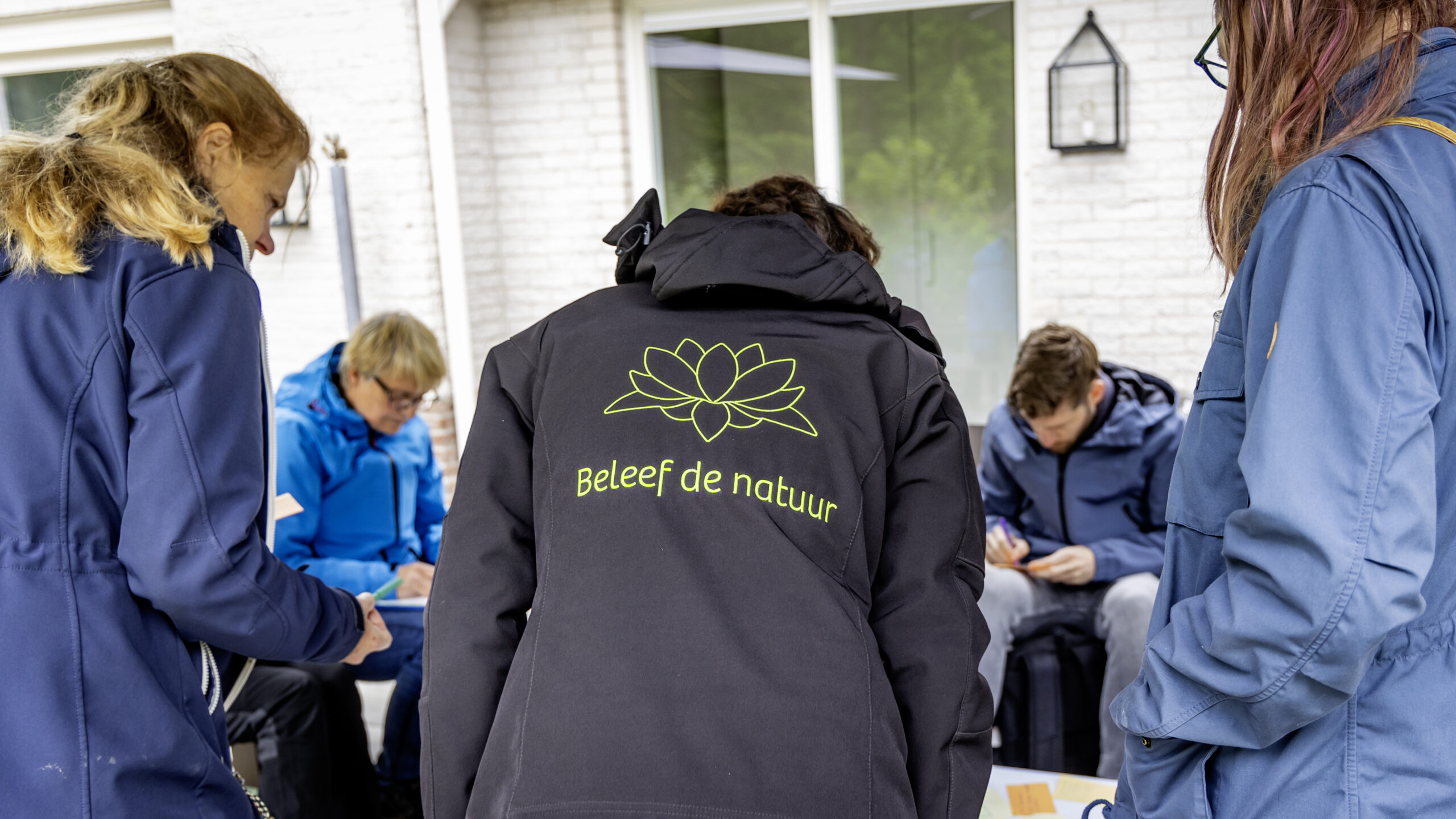 Groep mensen in blauwe jassen, één met "Beleef de natuur" logo, bezig met een activiteit buiten.