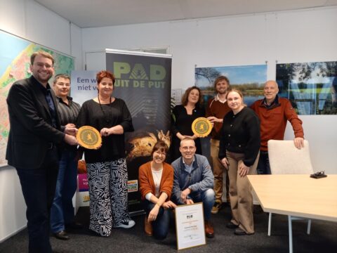 Een groep mensen poseert met een accolade in een kantoor met posters en een kaart.