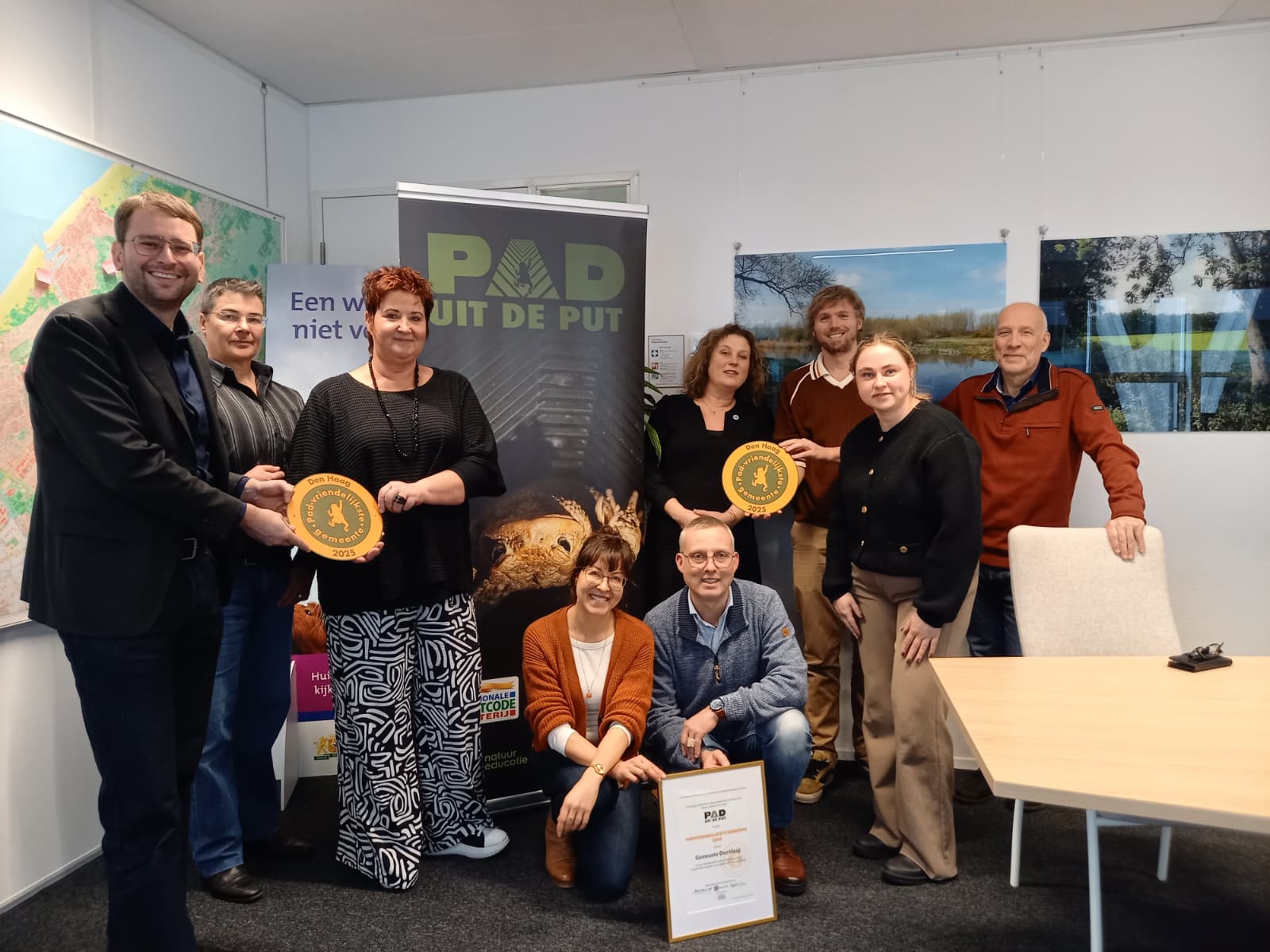 Een groep mensen poseert met een accolade in een kantoor met posters en een kaart.