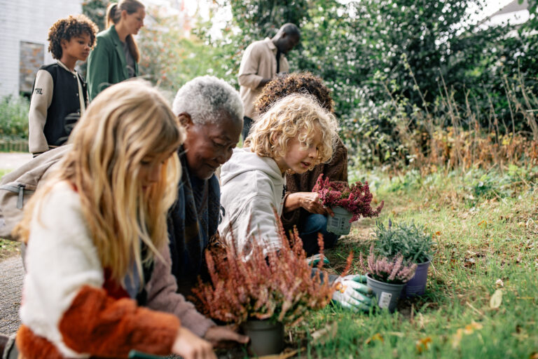 Groep mensen plant bloemen in een tuin, met focus op een jong kind en oudere dame vooraan.