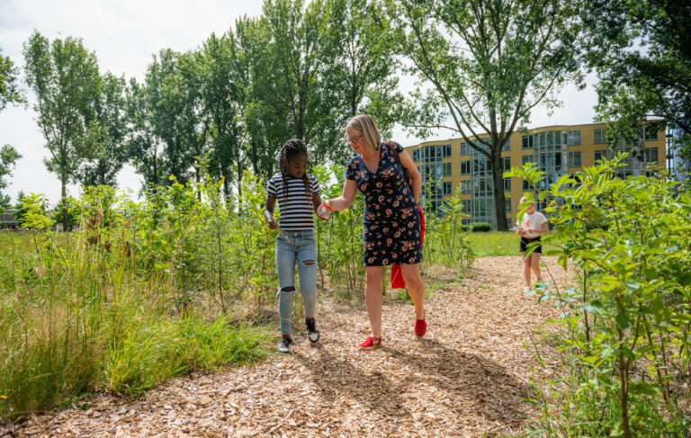 Vrouw en kind wandelen op een bospad, omringd door groen en bomen achter een appartementencomplex.