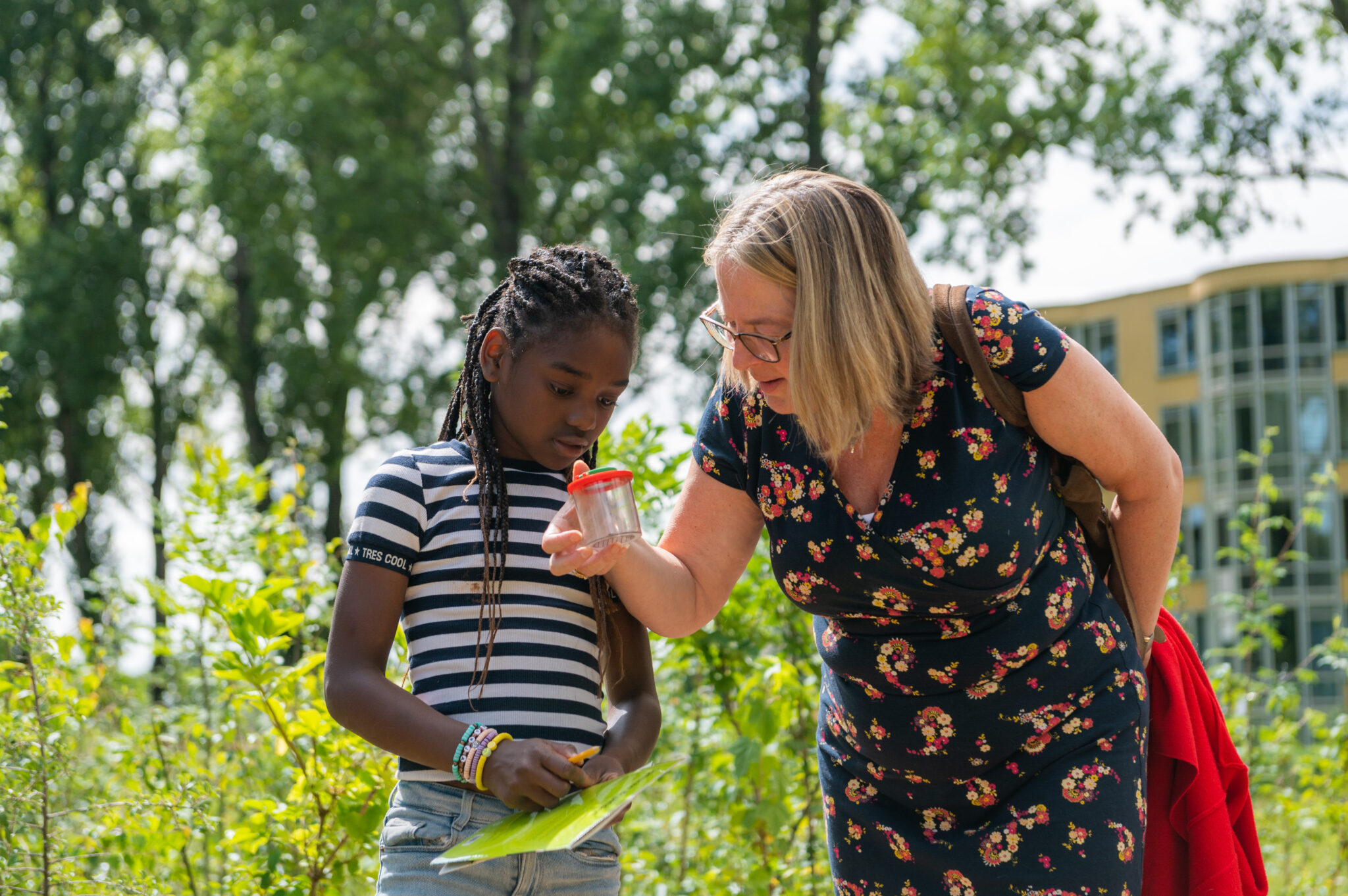 Kind en vrouw onderzoeken samen een insect in een potje in een groene buitenomgeving.