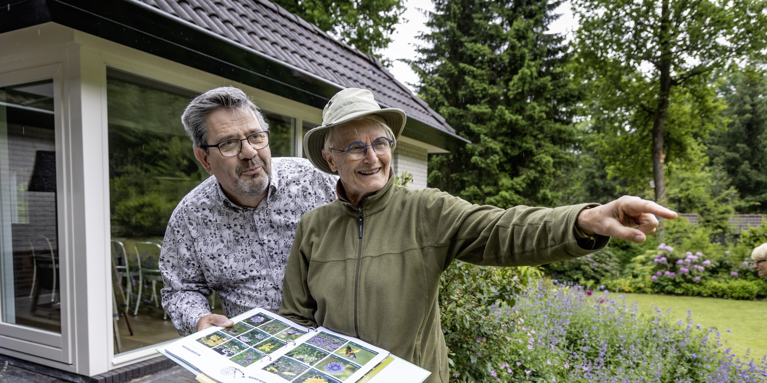 Twee mensen kijken naar planten in een tuin, met een open boek over flora.