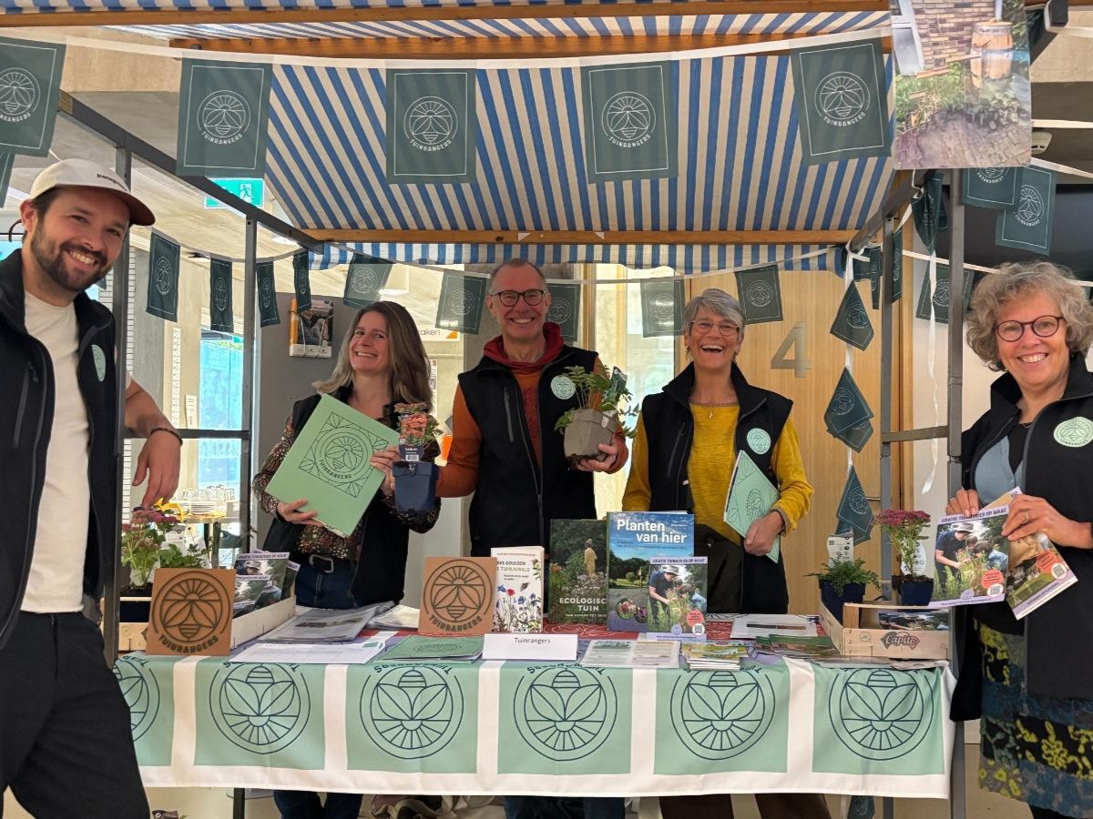 Groepsfoto bij een marktkraam met tuinposters en planten, bemand door vrolijke mensen.